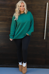 Woman model wearing a green oversized sweater with black leggings, white socks, and tan Uggs. The photo is taken outdoors against a dark brown door. 