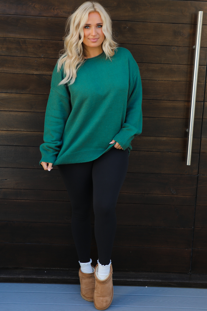 Woman model wearing a green oversized sweater with black leggings, white socks, and tan Uggs. The photo is taken outdoors against a dark brown door. 