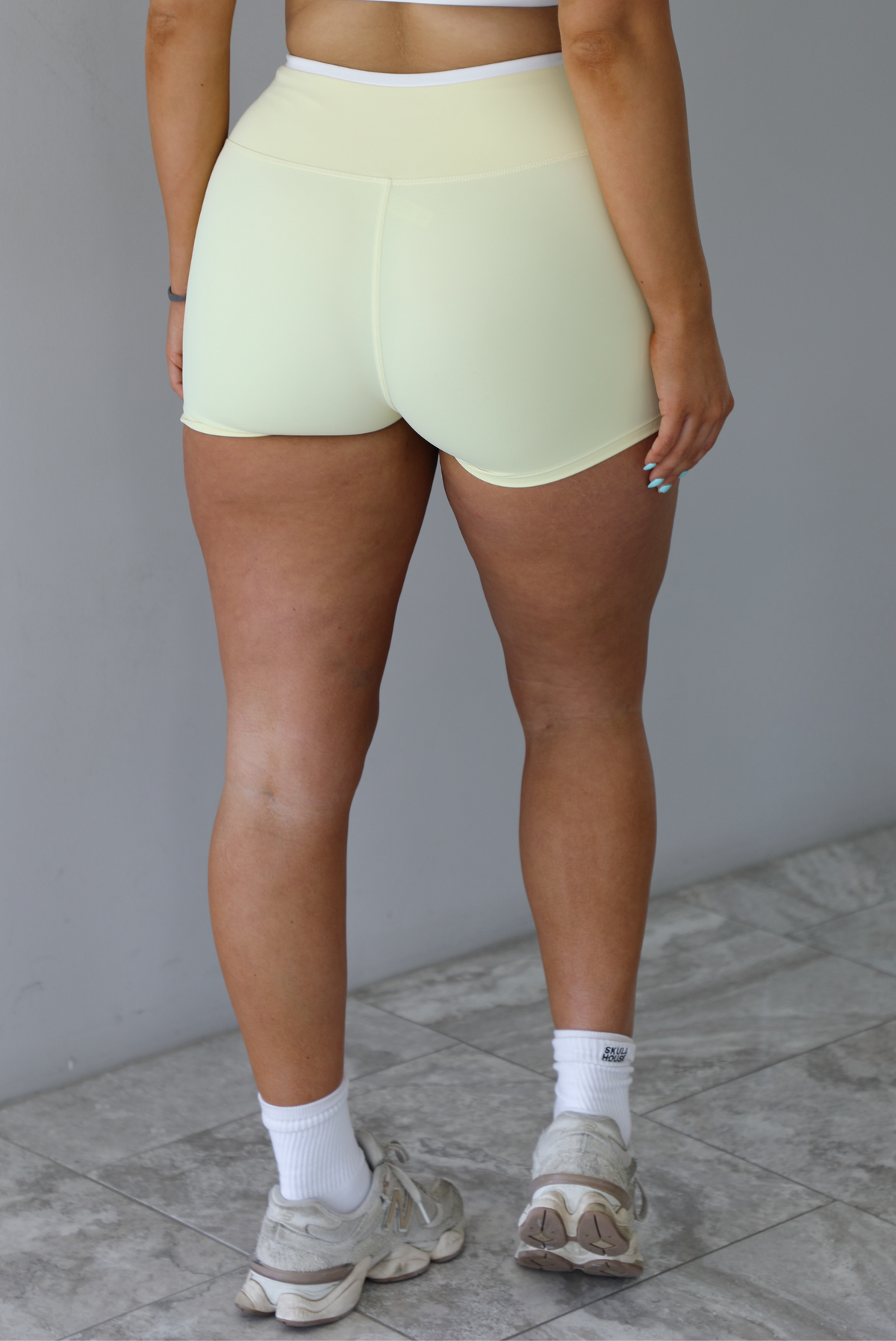 Woman wearing a yellow and white two piece athleisure set featuring a sports bra and biker shorts that have a layered look with the white peeking out from under the yellow. The photo is taken indoors against a white wall and shows a closeup of the back of the shorts. 