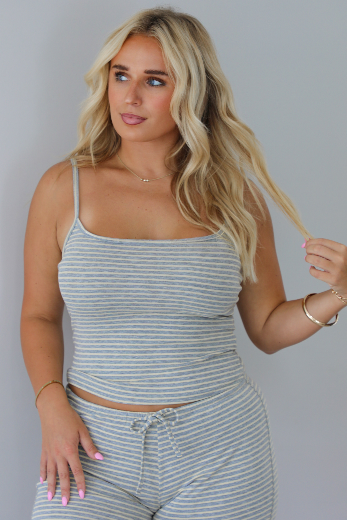 A blonde woman modeling a matching two-piece loungewear set consisting of a gray and pale yellow striped crop tank top and coordinating drawstring shorts, set indoors against a white wall. 