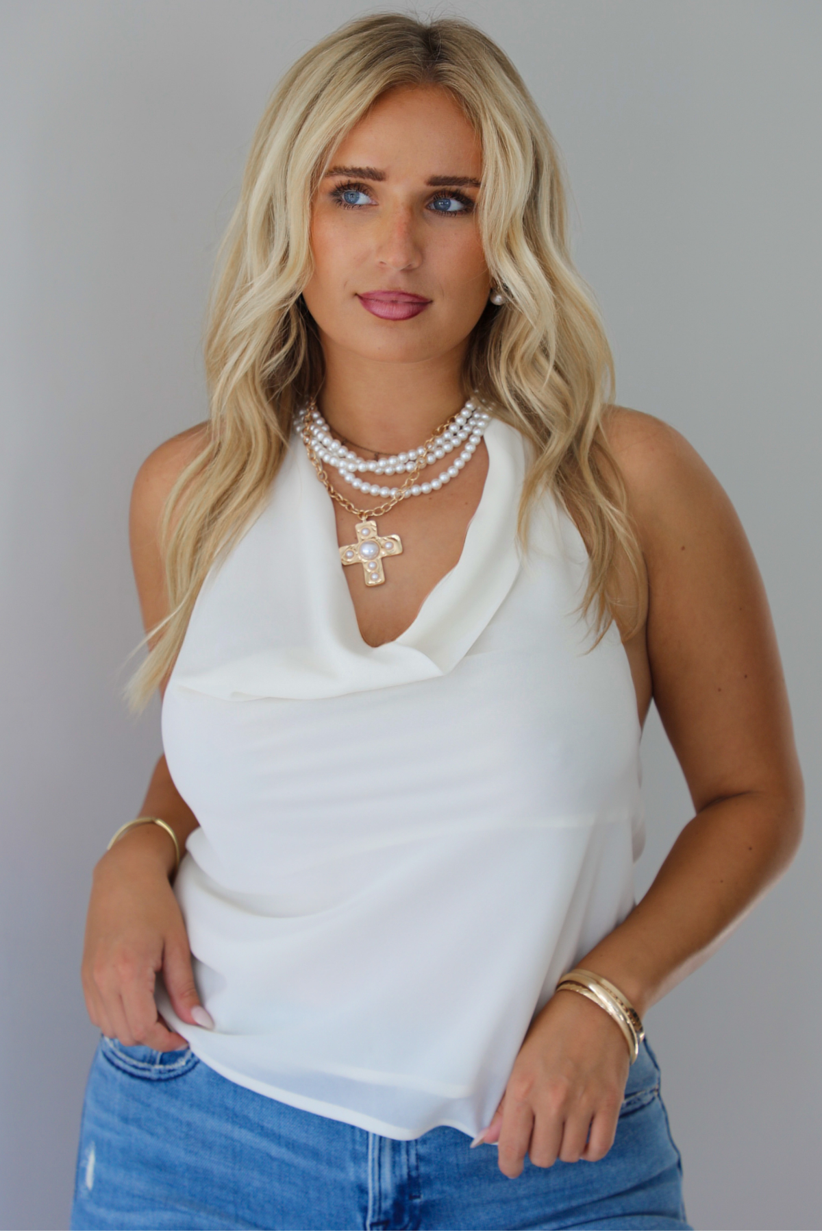 A woman modeling a cream sleeveless halter top and light-wash blue distressed flare jeans, standing indoors against a white wall.