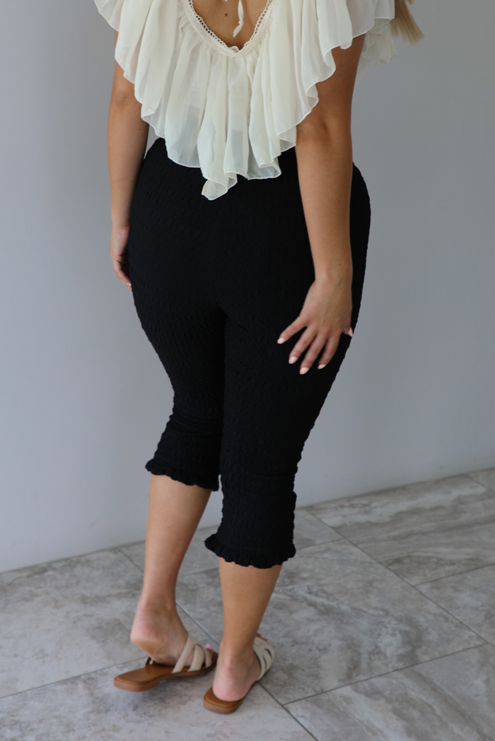 Closeup of a woman wearing high waisted black capri pants that have a textured feel to them and a slight ruffled hem indoors against a white wall with white marble flooring. The photo shows the back of the pants. 
