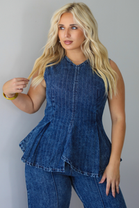 A woman wearing a matching two piece dark wash denim set with a pin striped design on it featuring a sleeveless peplum style top and high waisted denim jeans, standing indoors against a white wall.