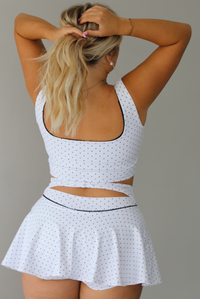 A woman shown from the back wearing a white two-piece athletic set with small black polka dots. The set includes a scoop-neck crop top with a matching ruffled mini skirt that has a cutout at the waist.