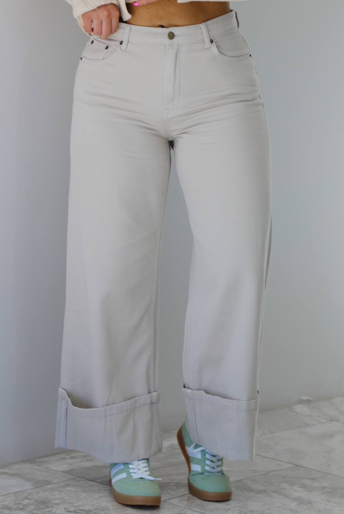 Woman wearing high waisted, sand colored denim pants that have a straight leg cut with a cuffed hem. The photo is taken indoors against a white wall with white marble flooring. 