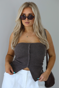 Woman wearing a strapless brown top with non-functional button closures in the front with  a split hem detail, and high waisted, flared hem white linen pants with a brown shoulder bag. The photo is taken indoors against a white wall and shows a closeup of the brown top. 