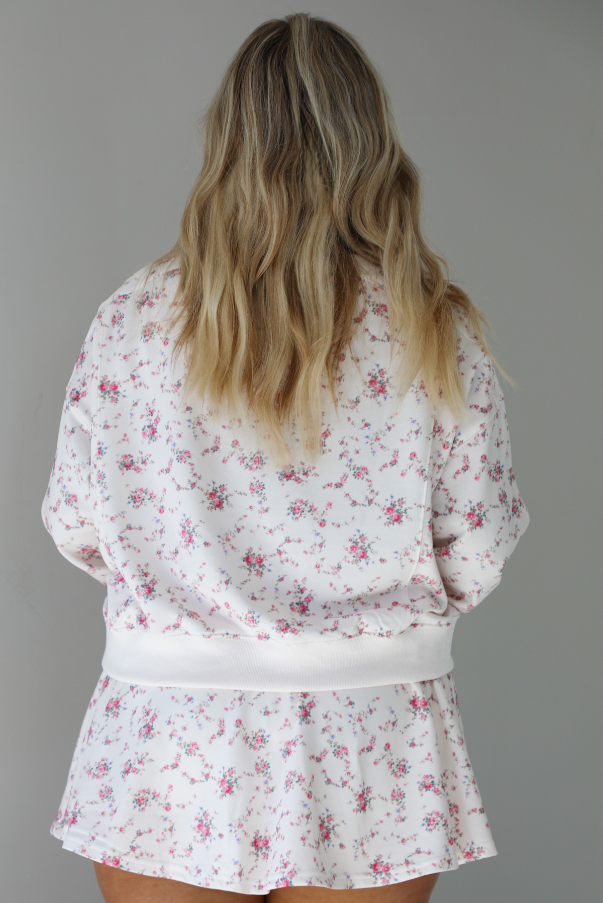 Woman wearing a pale pink two piece set that has a pink floral print pattern throughout. The set featuring an oversized long sleeved top and high waisted skirt. the photo shows the back of the set. The photo is taken indoors against a white wall.