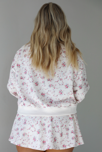 Woman wearing a pale pink two piece set that has a pink floral print pattern throughout. The set featuring an oversized long sleeved top and high waisted skirt. the photo shows the back of the set. The photo is taken indoors against a white wall.