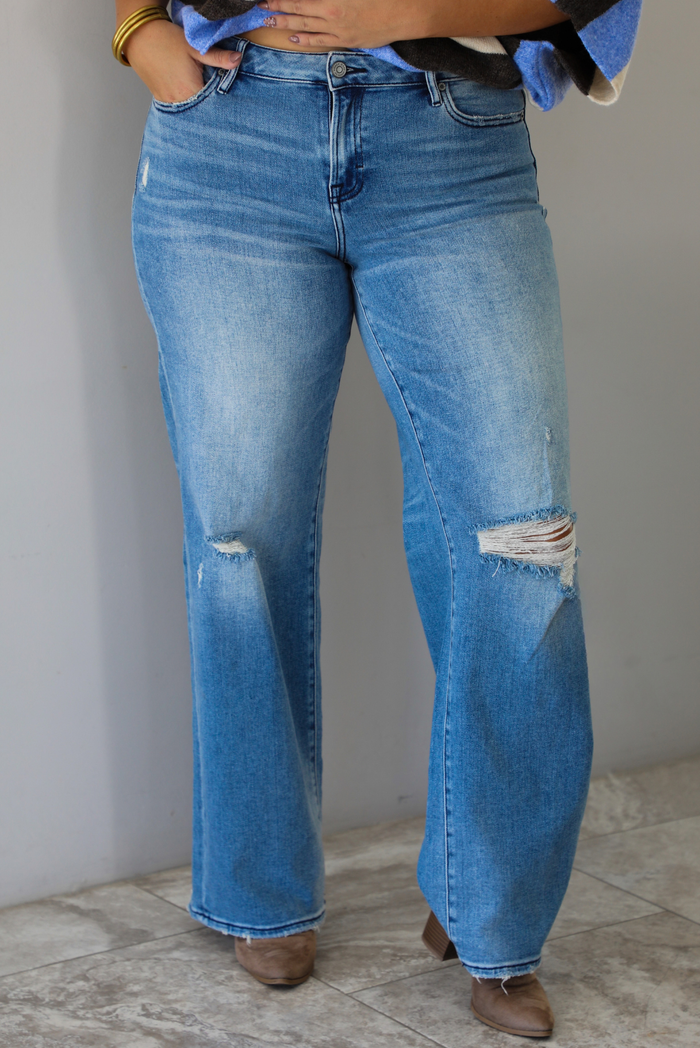 Closeup of a woman wearing mid-wash denim jeans with a mid-rise waistline, distressed details, and flared hems indoors against a white wall with white marble flooring. 