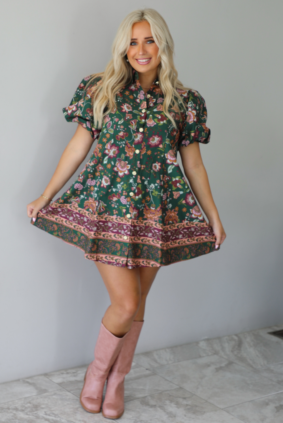 A woman wearing a dark green dress that has a mauve and multicolored floral print pattern throughout, has puffed sleeves, a collared neckline with functional gold button closures in the front, paired with muted pink boots, standing indoors against a white wall with white marble flooring. 