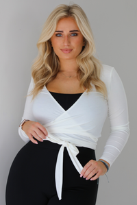 Woman wearing a white cropped wrap style fitted top over a black fitted jumpsuit. The photo shows a closeup of the wrap top. The photo is taken indoors against a white wall with white marble flooring. 