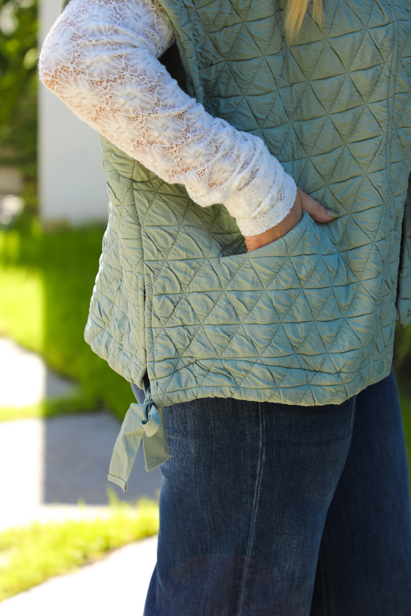 Close up of the pocket on the vest on a woman model wearing a sage colored oversized vest paired with a white lace long sleeved top and dark denim outdoors.  The vest has functional pockets on either side, functional button closures in the front, and an oversized fit. 