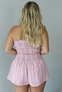 A woman wearing a pink and multi-colored vertically striped strapless romper, standing indoors against a white wall. The photo shows a closeup of the back of the romper. 