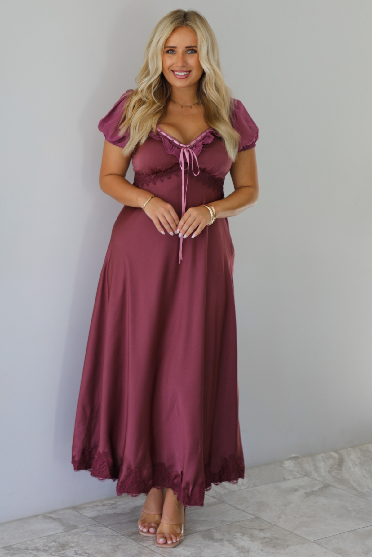 A woman modeling a long, burgundy maxi dress with short puff sleeves, a lace-up sweetheart neckline, and a flowing satin-like skirt, standing indoors against a white wall with white marble flooring. 