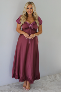 A woman modeling a long, burgundy maxi dress with short puff sleeves, a lace-up sweetheart neckline, and a flowing satin-like skirt, standing indoors against a white wall with white marble flooring. 