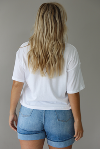 Woman wearing a white cropped boxy tee with a pink, red, and yellow bikini graphic on the front with a pair of medium wash denim shorts indoors against a white wall. The photo shows the back of the tee.