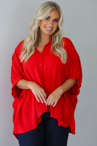 A woman wearing a bright red oversized short-sleeve blouse with functional button closures in the front and dark wide-leg jeans, standing indoors against a white wall.
