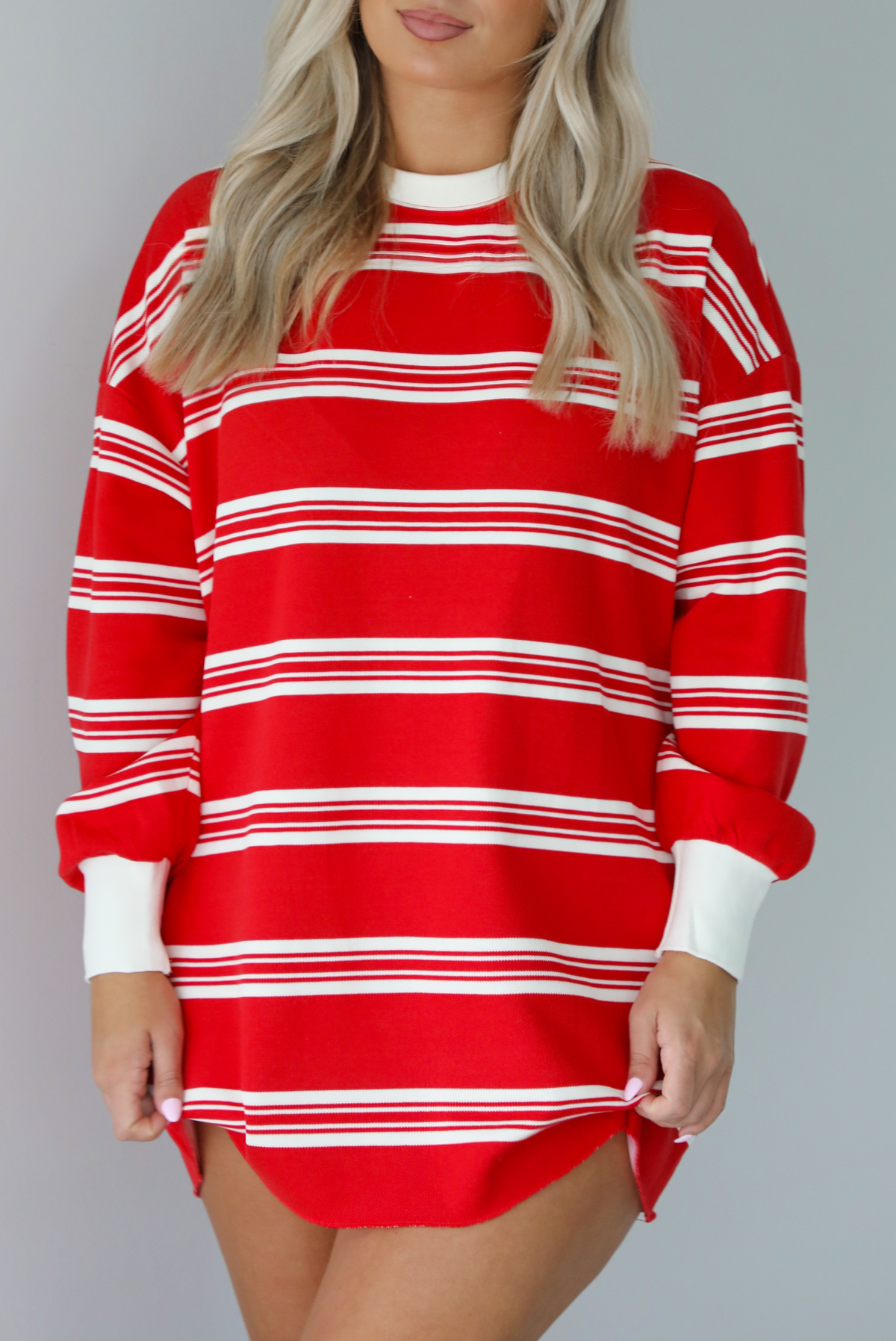 Woman wearing a red with white stripes long sleeved t-shirt dress. The photo is taken indoors against a white wall.