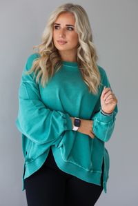 Woman wearing a teal oversized sweater with black leggings that have a slightly flared hem with split hem details in the front. The photo shows a closeup of the sweater and has a white background. 