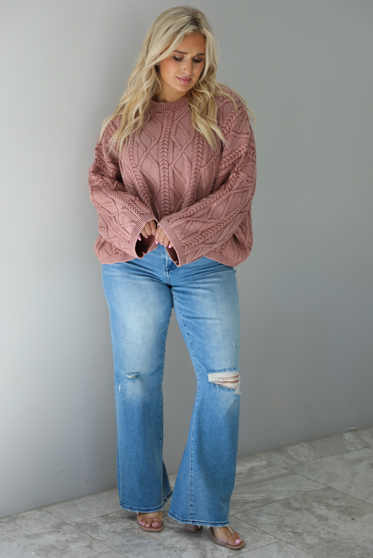 A woman wearing a mulberry oversized sweater paired with a pair of medium wash distressed denim, standing indoors against a white wall with white marble flooring. 
