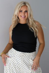 A woman wearing a black high neck tank paired with sand white pants with a black polka dot print pattern throughout, standing indoors against a white wall.