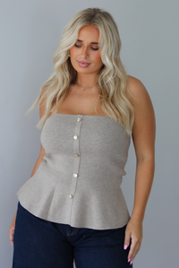A woman wearing a beige strapless button-front peplum top and dark denim wide-leg jeans, standing indoors against a white wall.