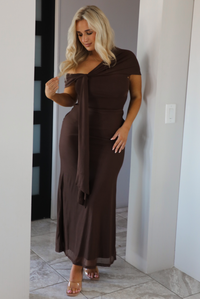 Woman model wearing a chocolate brown formal style dress that's fitted at the bodice, has asymmetrical sleeves, and a mermaid style skirt that's fitted at the hips and flared from the bottom of the high to the hem. The photo is taken inside a modern home in an archway.