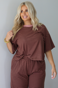 A woman wearing a brown and pink striped two piece set featuring a slightly cropped tee and high waisted pants, with the top slightly tucked into the pants, standing indoors against a white wall. The photo shows a closeup of the top, showing the pink trim neckline. 