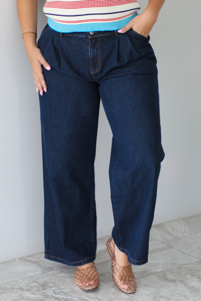 A person wearing dark blue wide-leg denim jeans, paired with a striped top and brown woven flat shoes, standing indoors against a white wall with white marble flooring.
