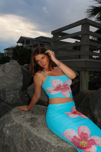 A woman wearing a bright turquoise two-piece set with large pink and white floral prints, posing on a large gray rock at a beach with wooden deck and houses in the background.