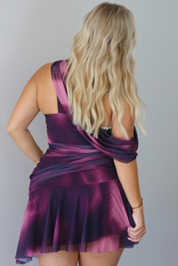 A woman modeling a short, one-shoulder mini dress in a deep purple with light purple design, paired with black ankle boots, standing indoors against a white wall. Th photo shows the back of the dress. 