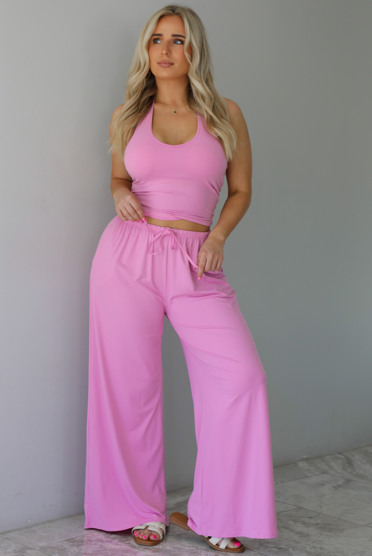 Woman wearing a two pieced pink set featuring a halter top and comfy pants with a flared hem detail. The photo is taken indoors against a white wall with white marble flooring. 