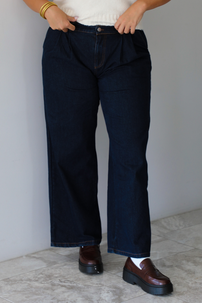 A woman wearing dark denim wide-leg jeans, a cream knit top, and brown leather shoes, standing indoors against a white wall with white marble flooring. 