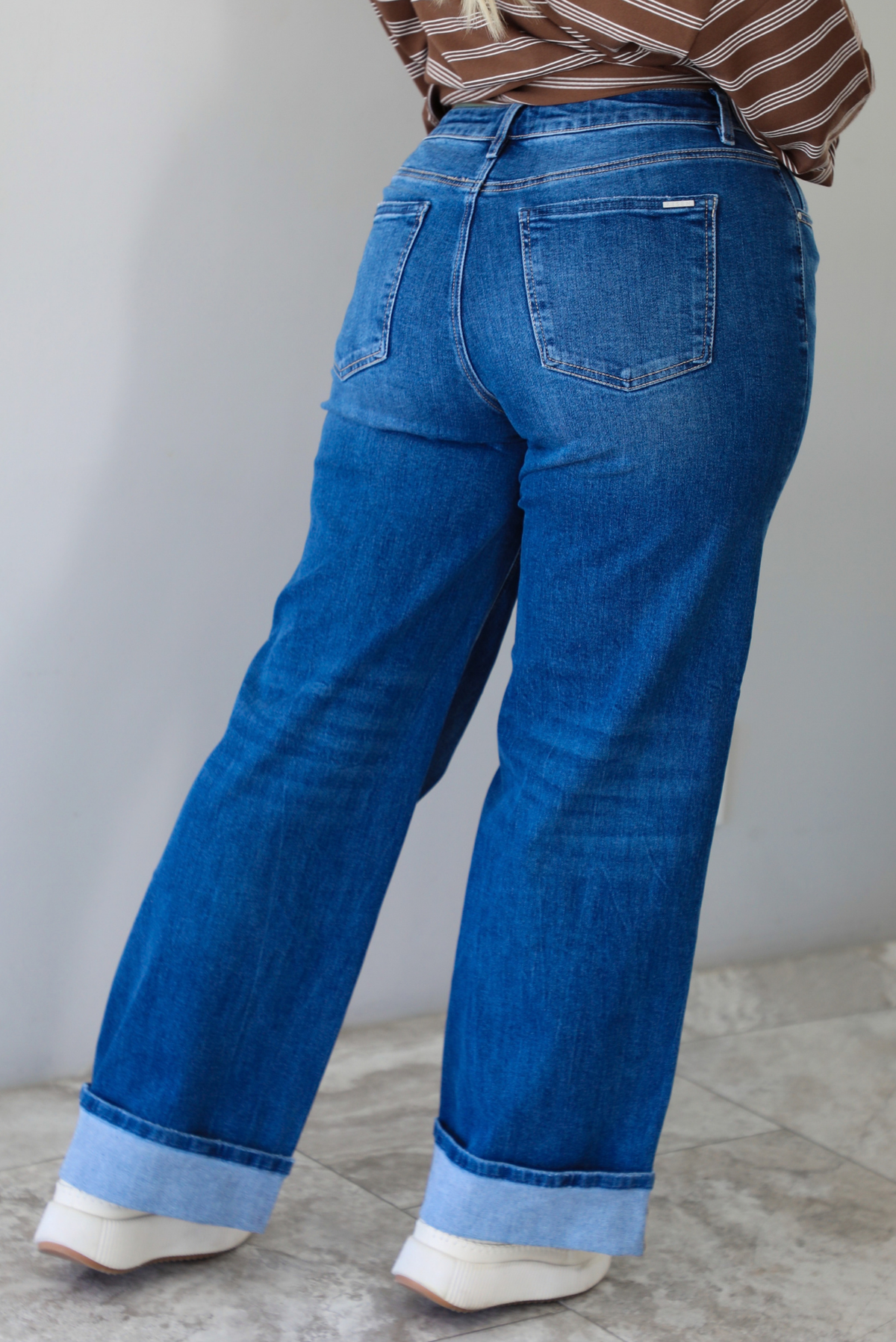 The closeup of a woman from behind  wearing dark wash denim jeans with a brown and white striped top and cream colored sneakers, standing indoors against a white wall with white marble flooring. 