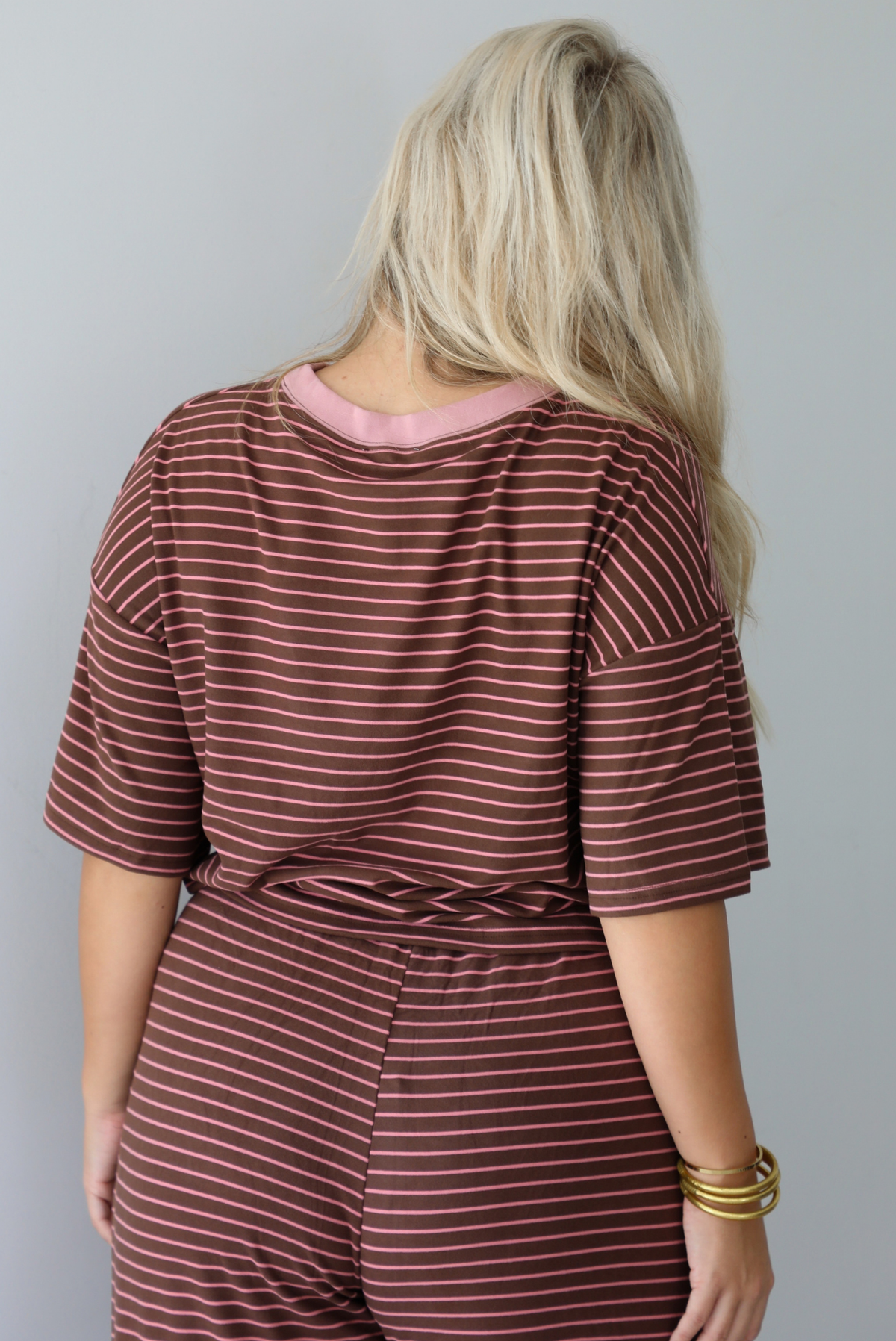 A woman shown from behind wearing a brown and pink striped two piece set featuring a slightly cropped tee and high waisted pants, with the top slightly tucked into the pants, standing indoors against a white wall. The photo shows a closeup of the back of the top.