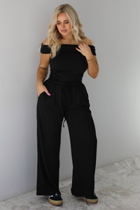 Woman wearing a two piece set featuring an off the shoulder top and high waisted pants. The photo is taken indoors against a white wall with white marble flooring. 