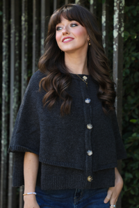 Close up of a woman model outdoors wearing a charcoal grey colored cape cardigan with functional button closures in the front and dark wash denim. 