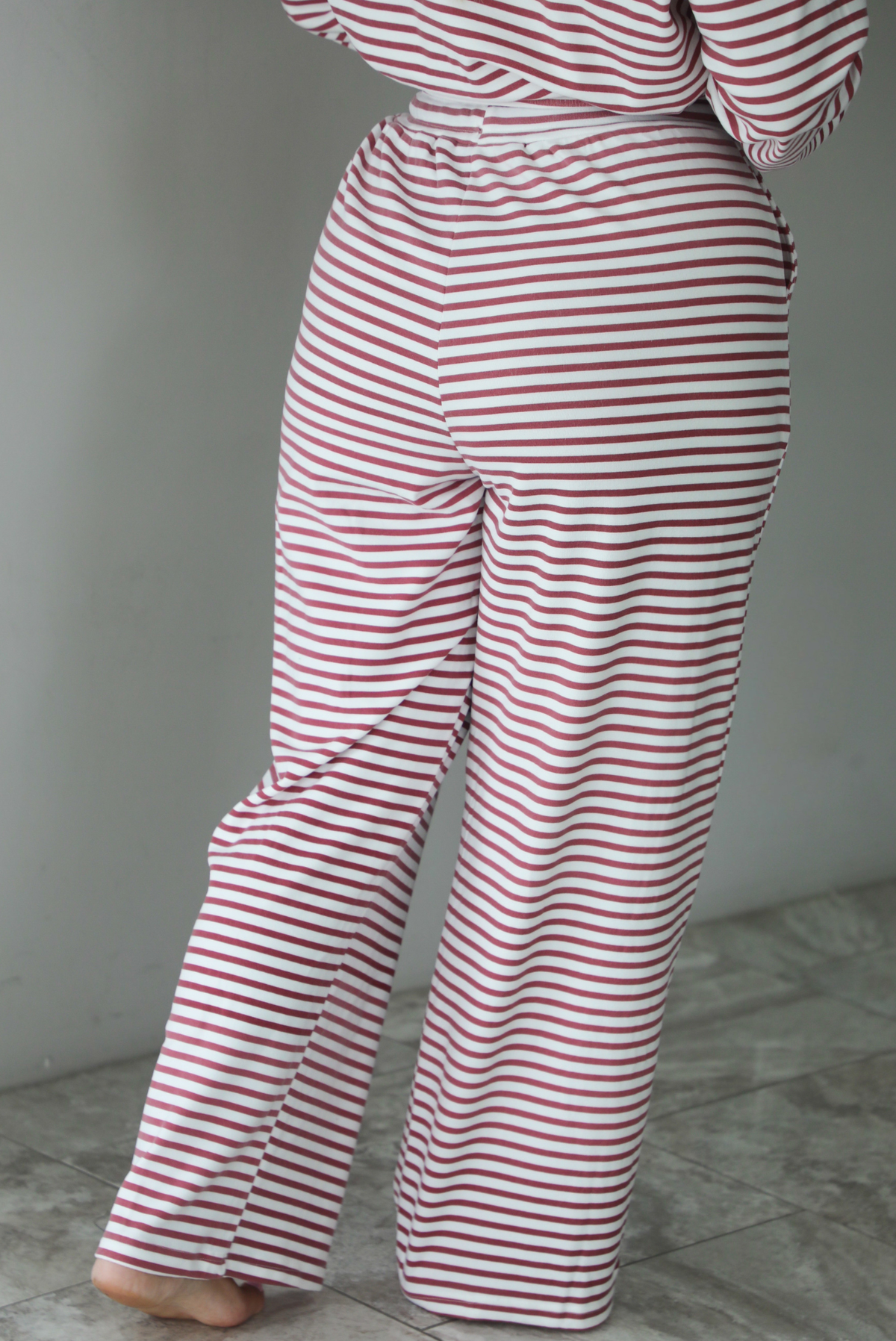 A woman shown from behind wearing a matching two piece set featuring a long sleeved top and high waisted wide leg pants with a berry and white striped pattern throughout, standing indoors against a white wall with white marble flooring. The photo shows a closeup of the back of the pants. 