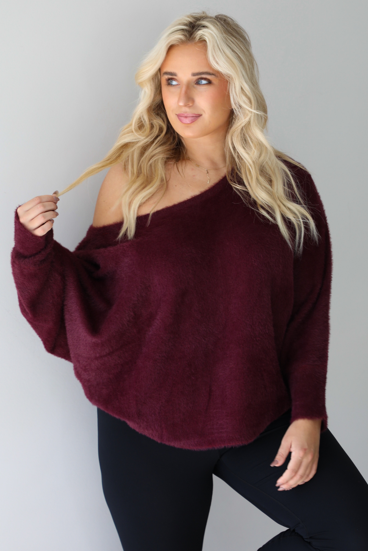 A woman wearing a maroon oversized long sleeved sweater, worn off the shoulder, paired with black leggings, indoors against a white wall.