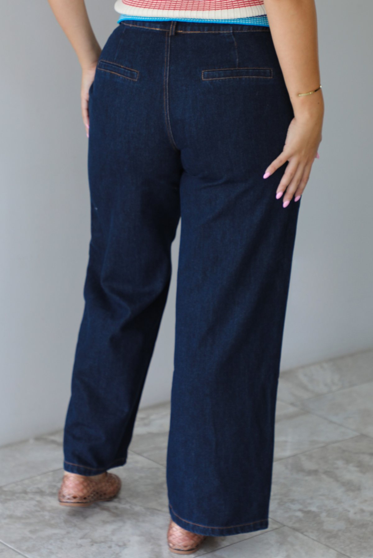 A person shown from behind wearing dark blue wide-leg denim jeans, paired with a striped top and brown woven flat shoes, standing indoors against a white wall with white marble flooring.