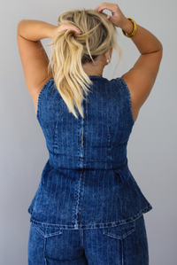A woman shown from behind wearing a matching two piece dark wash denim set with a pin striped design on it featuring a sleeveless peplum style top and high waisted denim jeans, standing indoors against a white wall. The photo focuses on the back of the top, which has a zipper closure. 