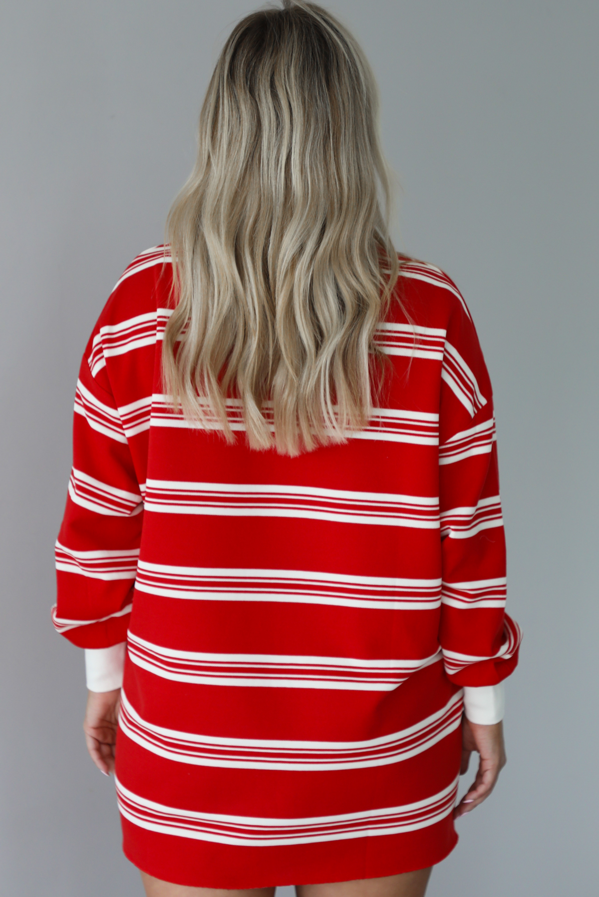 Woman wearing a red with white stripes long sleeved t-shirt dress. The photo is taken indoors against a white wall and shows the back of the dress. 