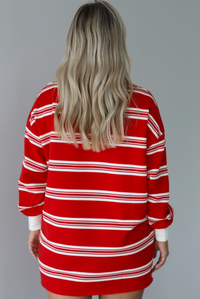 Woman wearing a red with white stripes long sleeved t-shirt dress. The photo is taken indoors against a white wall and shows the back of the dress. 