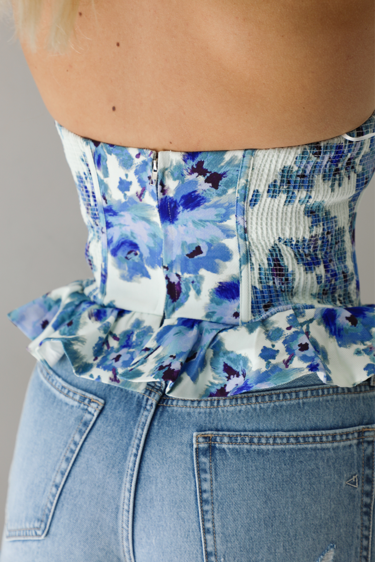 Woman wearing a blue and white strapless corset style top with a ruffled hem paired with light wash denim shorts. The photo is taken indoors against a white wall. The photo shows a closeup of the back of the top, which shows smocked detailing on the sides and a zipper closure in the back.