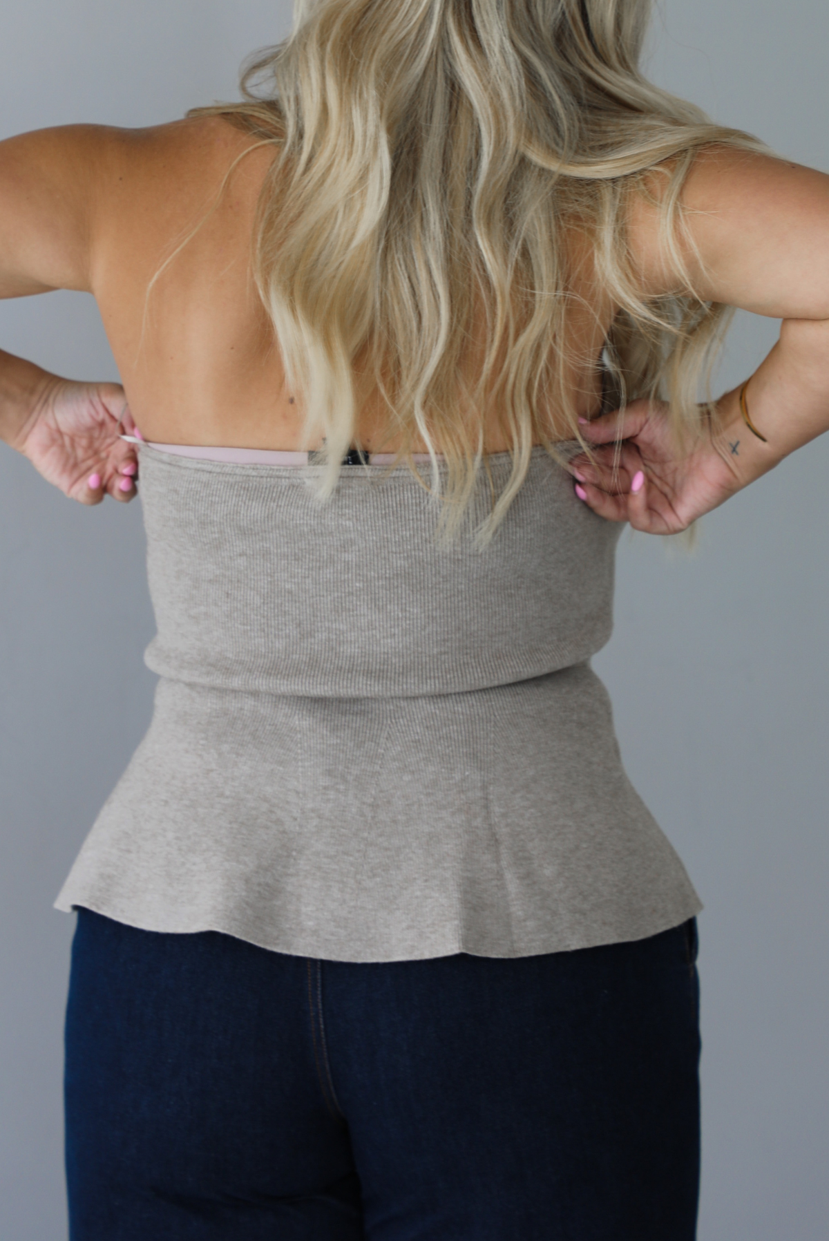 A woman shown from behind wearing a beige strapless button-front peplum top and dark denim wide-leg jeans, standing indoors against a white wall.