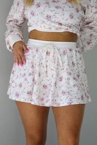 Woman wearing a pale pink two piece set that has a pink floral print pattern throughout. The set featuring an oversized long sleeved top and high waisted skirt. The photo shows a closeup of the skirt, which has an elastic drawstring waistline. The photo is taken indoors against a white wall.