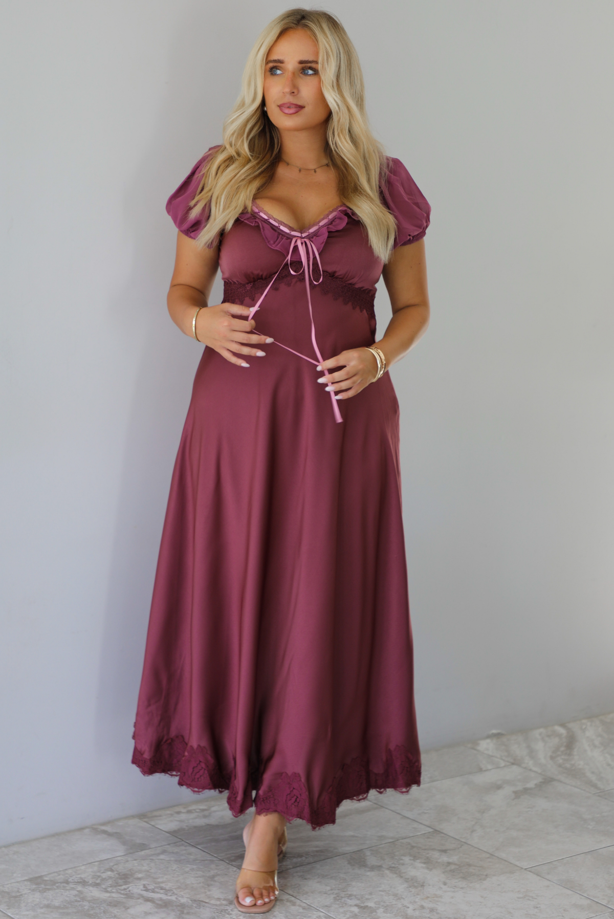 A woman modeling a long, burgundy maxi dress with short puff sleeves, a lace-up sweetheart neckline, and a flowing satin-like skirt, standing indoors against a white wall with white marble flooring. 