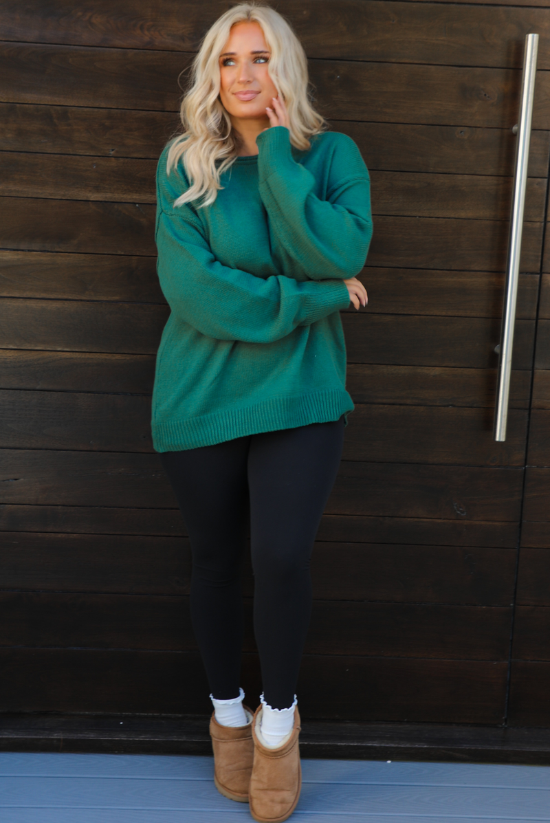 Woman model wearing a green oversized sweater with black leggings, white socks, and tan Uggs. The photo is taken outdoors against a dark brown door. 