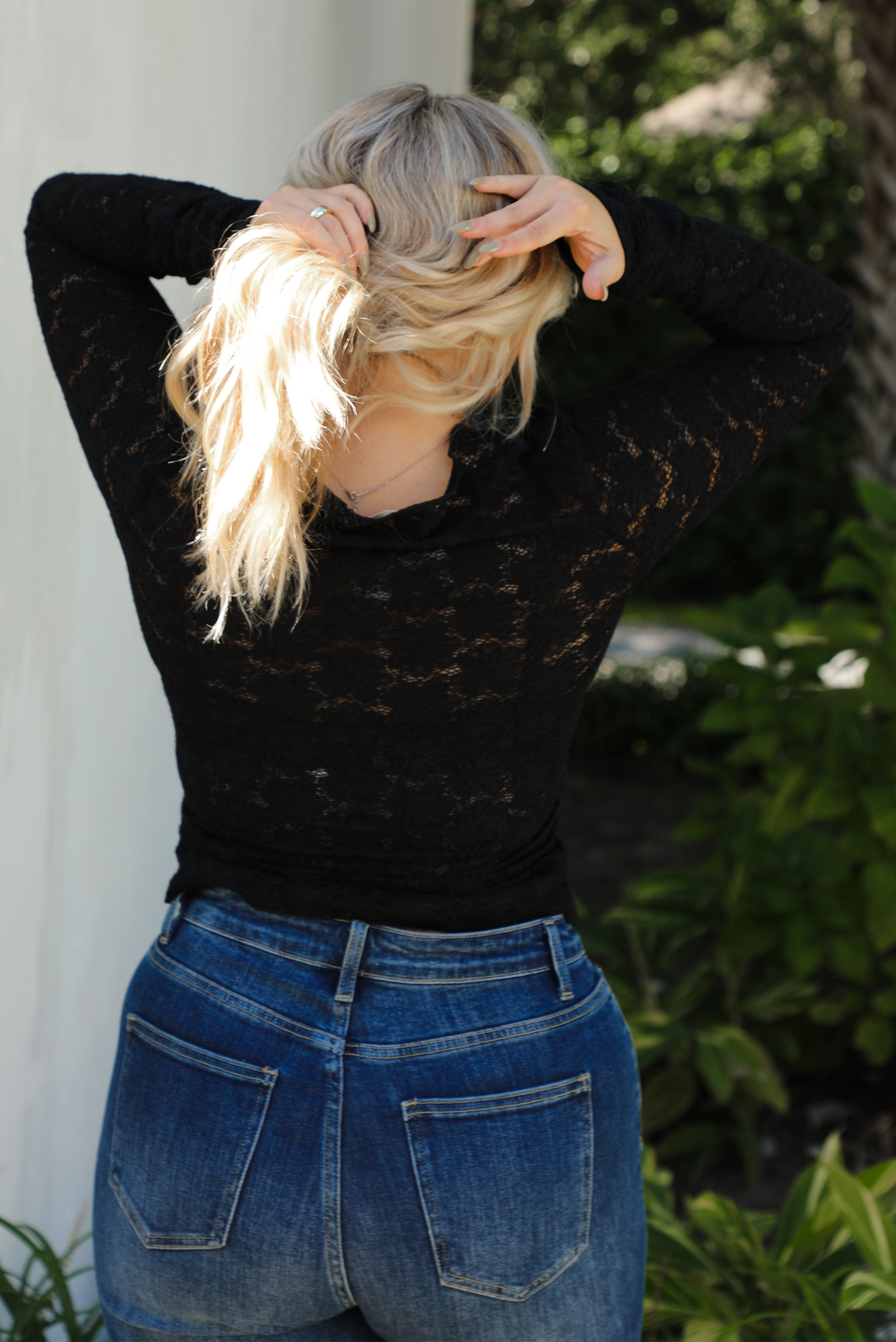 Close up of the back of a model wearing a black sheer lace top with a floral pattern with dark denim jeans outdoors.
