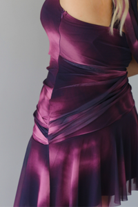 A woman modeling a short, one-shoulder mini dress in a deep purple with light purple design, paired with black ankle boots, standing indoors against a white wall. The photo shows the side of the dress, focusing on the zipper closure.